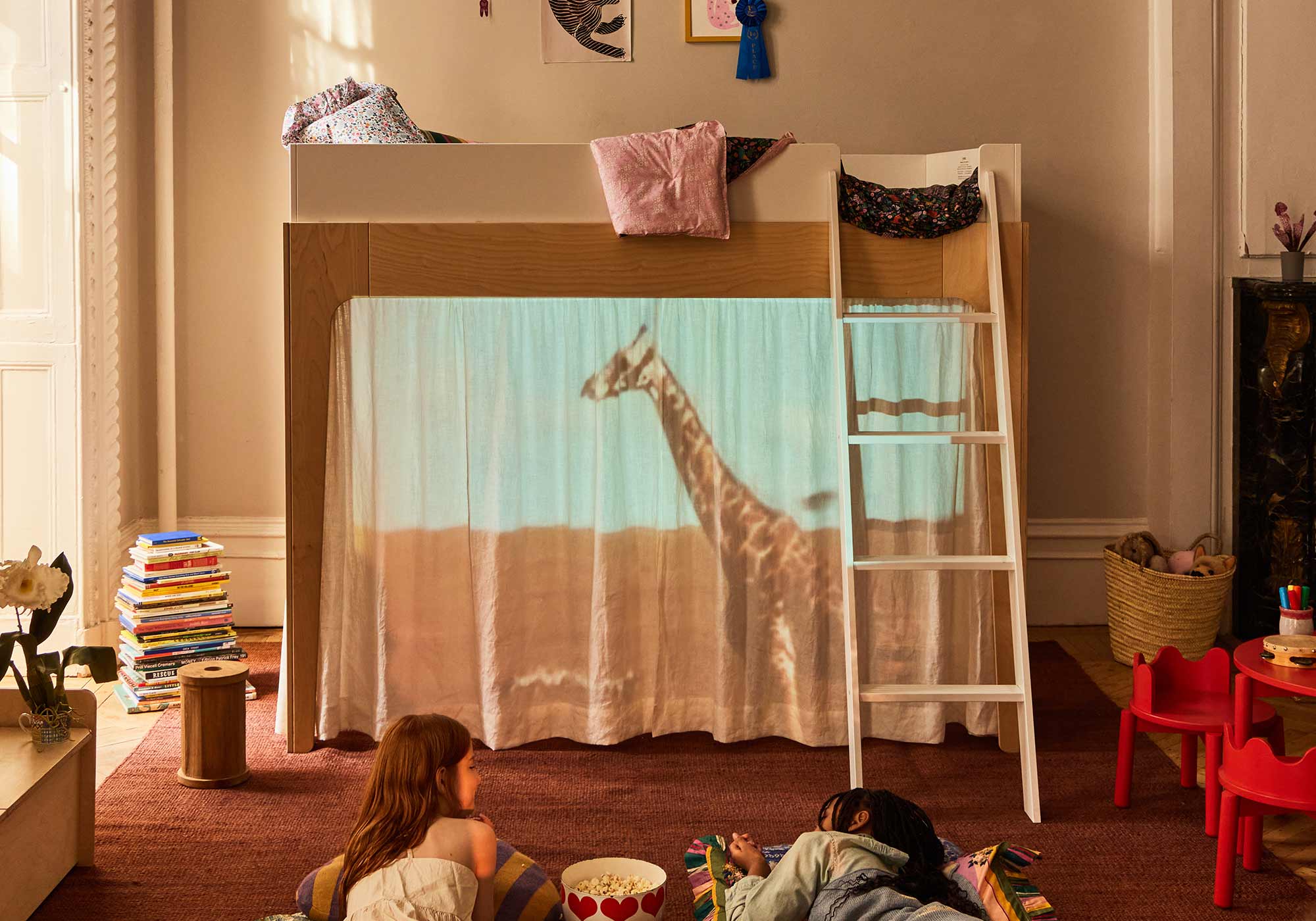Two children watch a giraffe projected on a curtain below a loft bed in a cozy, book-filled room.