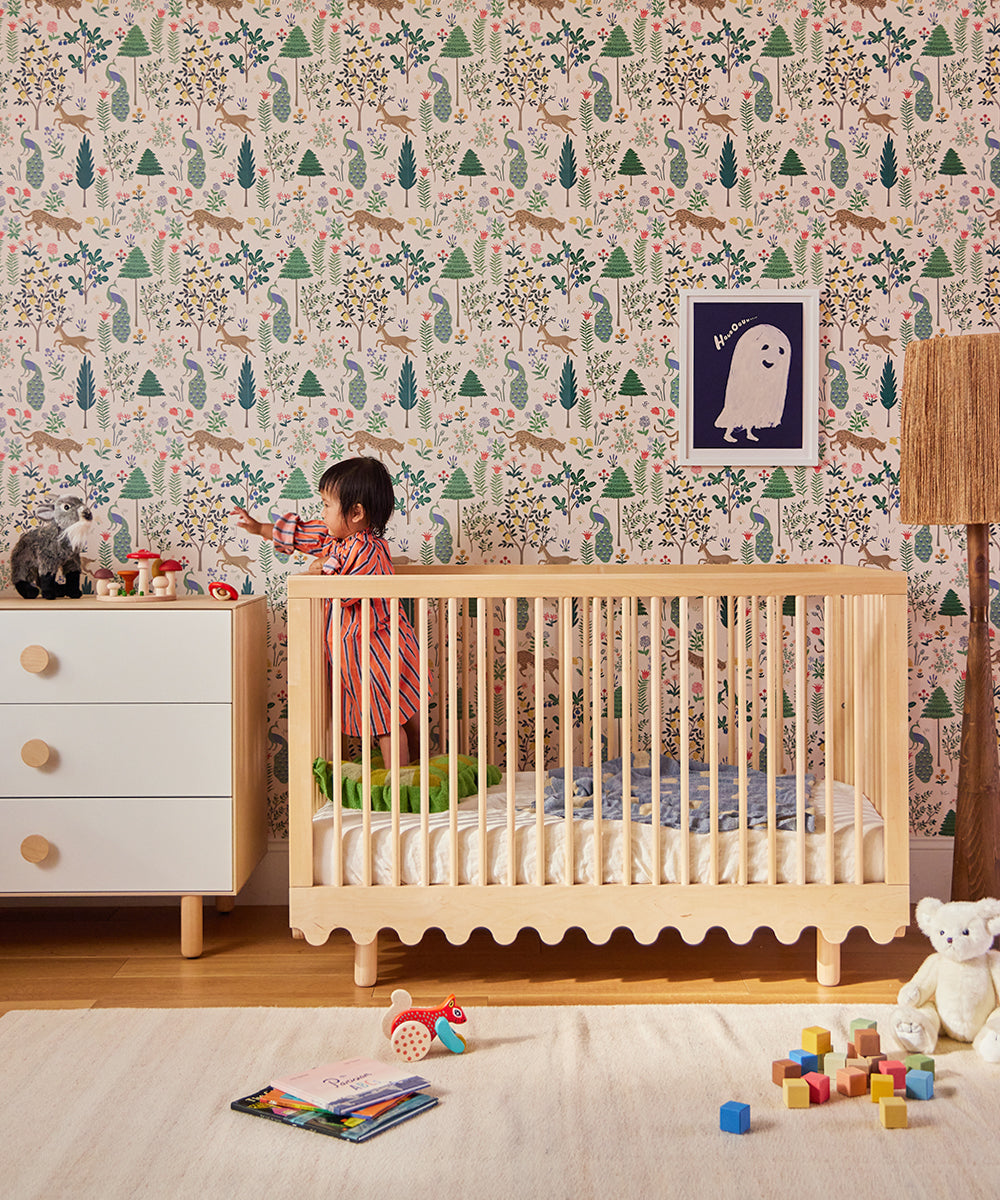 A toddler stands by a dresser in a colorful nursery with a crib, toys, animal wallpaper, and the Le Baby Bird Rattle.