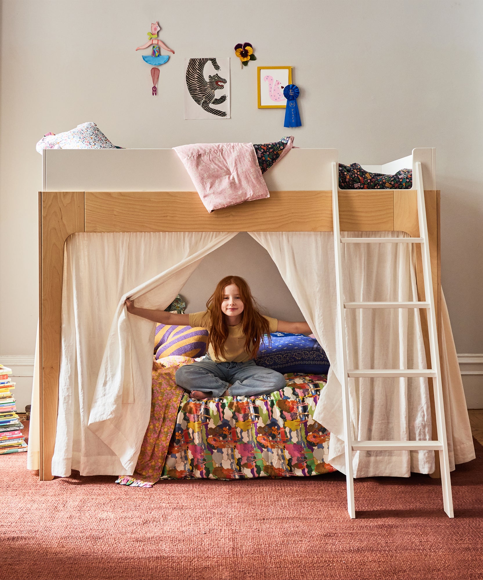 A red-haired girl smiles, sitting cross-legged on a colorful bed with Perch Bunk Bed Curtains beneath a loft and artwork above.