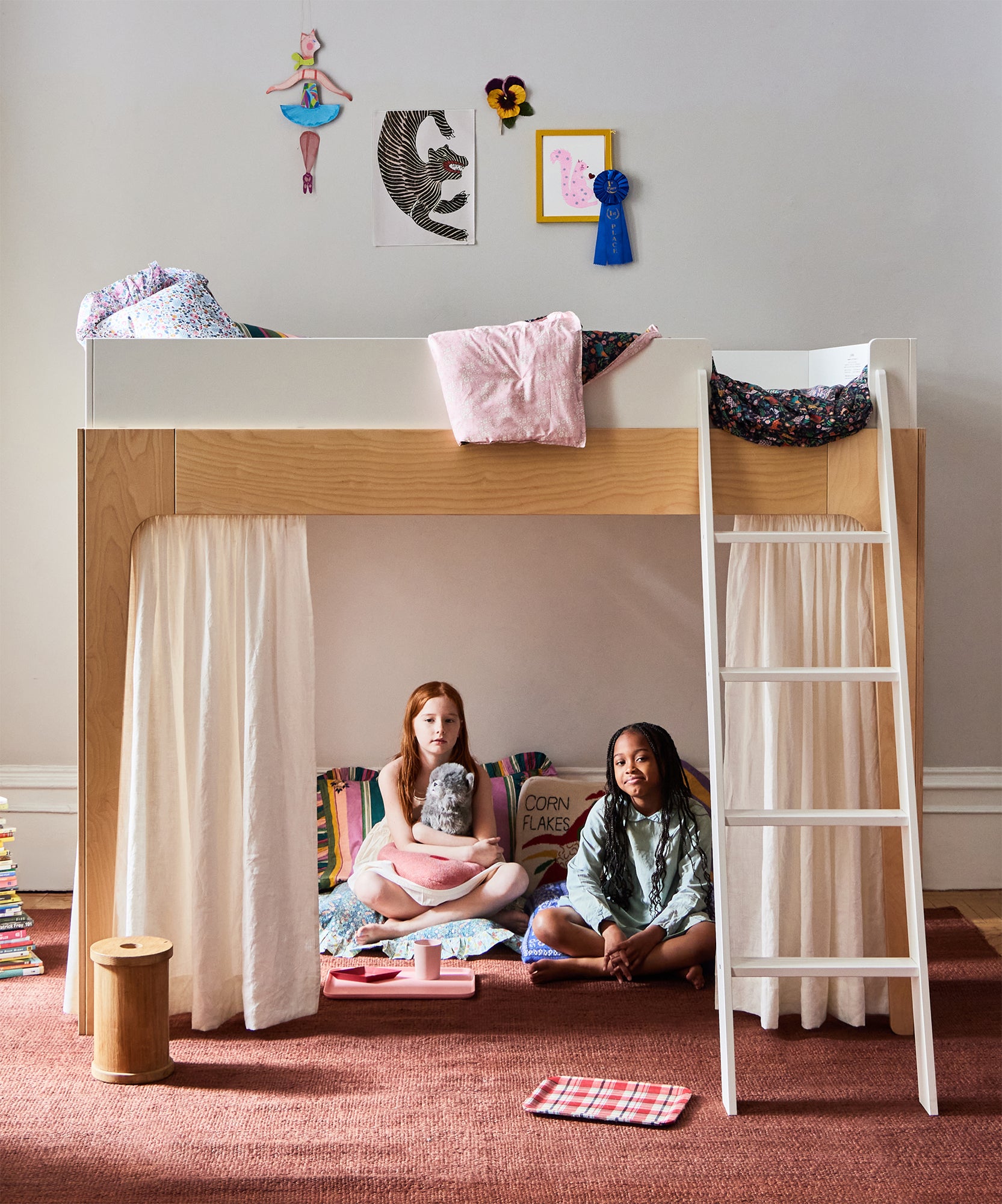 Two girls sit under a Perch Bunk Bed with Curtains, surrounded by pillows, stuffed animals, and books on a cozy rug in a child's bedroom.