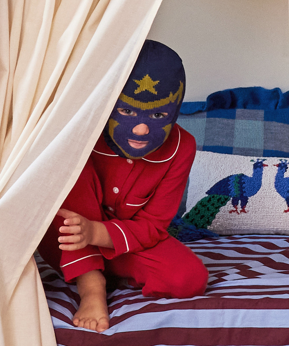 A child peeks out from behind Perch Bunk Bed Curtains, sitting on a bed with peacock pillows, dressed in red pajamas and a blue mask.