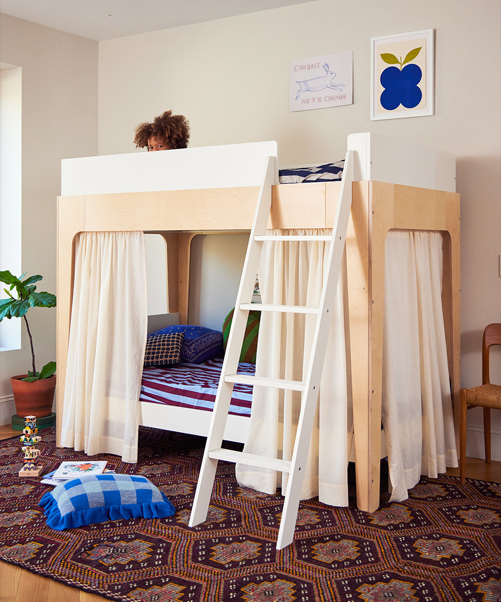 A child peeks from the top bunk of a modern wooden bed with a ladder, striped bedding, and Perch Bunk Bed Curtains.