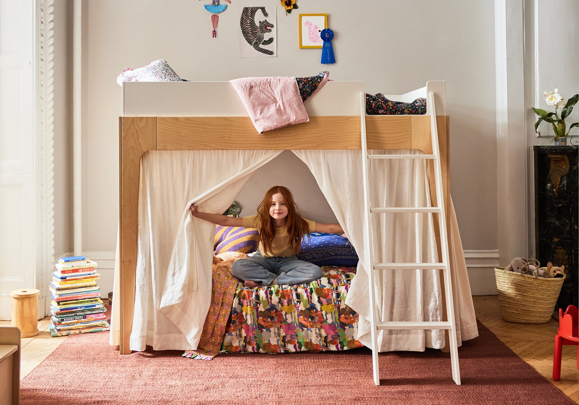 Smiling child sits inside a cozy bunk bed tent with books stacked and artwork on the wall.
