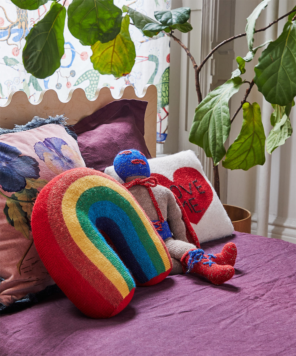 The Rainbow Pillow adds color to a purple bedspread with a knit doll, leafy plant, and floral pillow nearby.