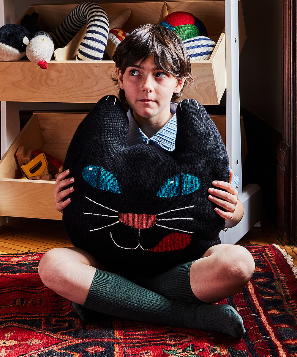 A child sits on a rug, holding the Black Cat Pillow, with toys stored in wooden bins in the background.