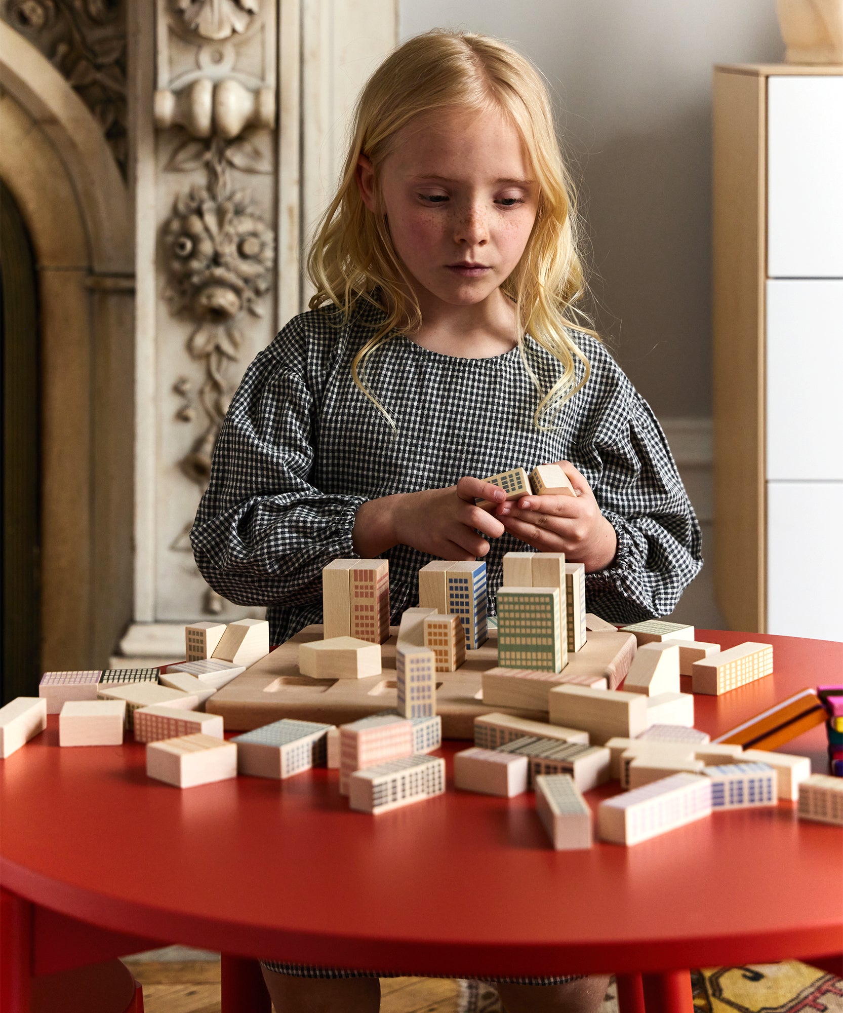 A young girl focuses intently as she builds a small city with Le Manhattan Skyline Blocks at a red table.