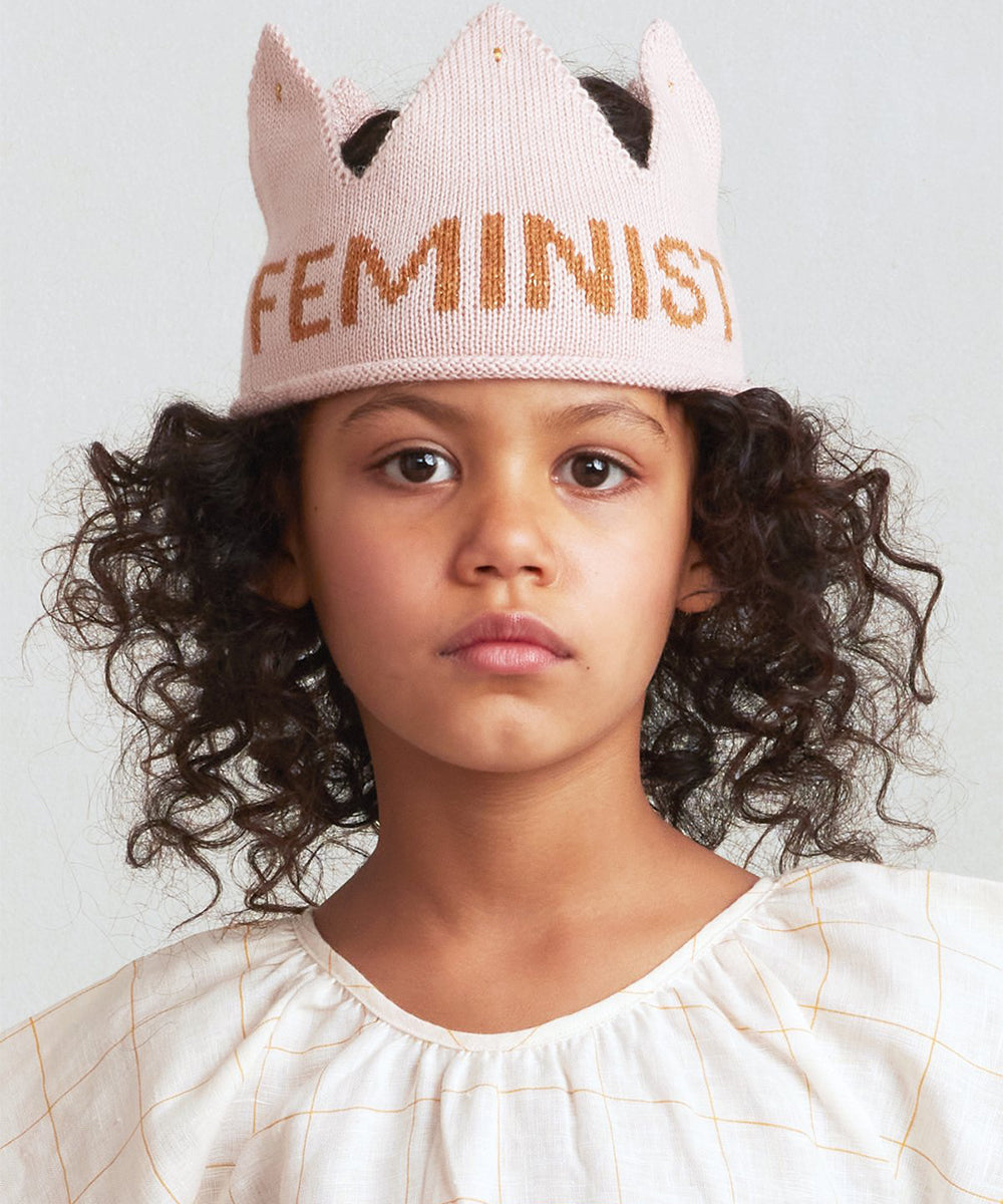 A child with curly hair, wearing a "Feminist Crown" and a white blouse, stands against a neutral background.